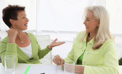 Zwei Frauen in grünen Oberteilen sitzen an einem Tisch mit Gläsern und sprechen miteinander.