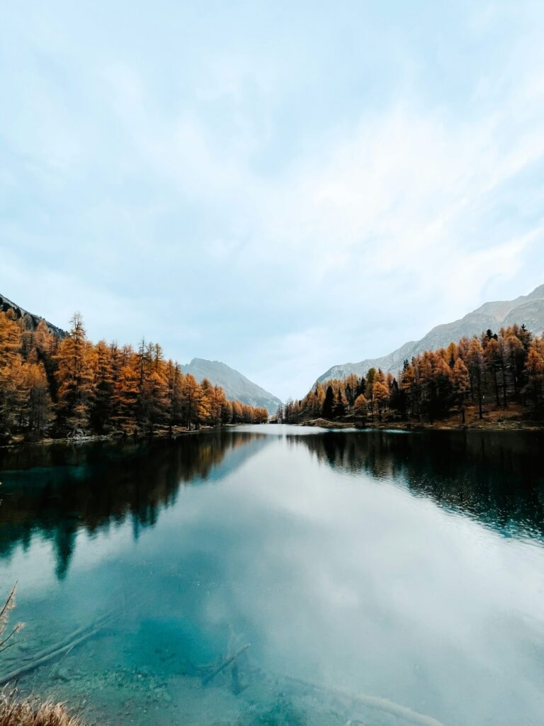 Bergsee mit spiegelglatter Wasseroberfläche, umgeben von herbstlich gefärbten Nadelbäumen und Bergen im Hintergrund.