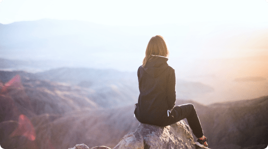 Person mit braunen Haaren sitzt auf einem Felsen und blickt auf eine neblige Berglandschaft bei Sonnenuntergang.
