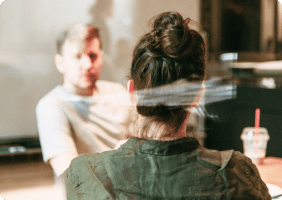 Rückansicht einer Person mit zusammengebundenem Haar, die einem anderen gegenüber sitzt, auf einem Tisch steht ein Getränk mit rotem Strohhalm.