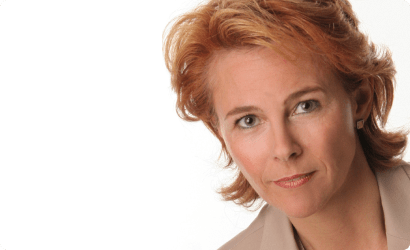 Person mit halblangen, gewellten, rotblonden Haaren, trägt ein helles Hemd und einen beigen Blazer, vor weißem Hintergrund.