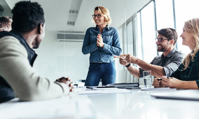 Drei Personen an einem Tisch, eine steht mit verschränkten Händen, zwei sitzen, einer zeigt mit ausgestrecktem Arm nach vorne.